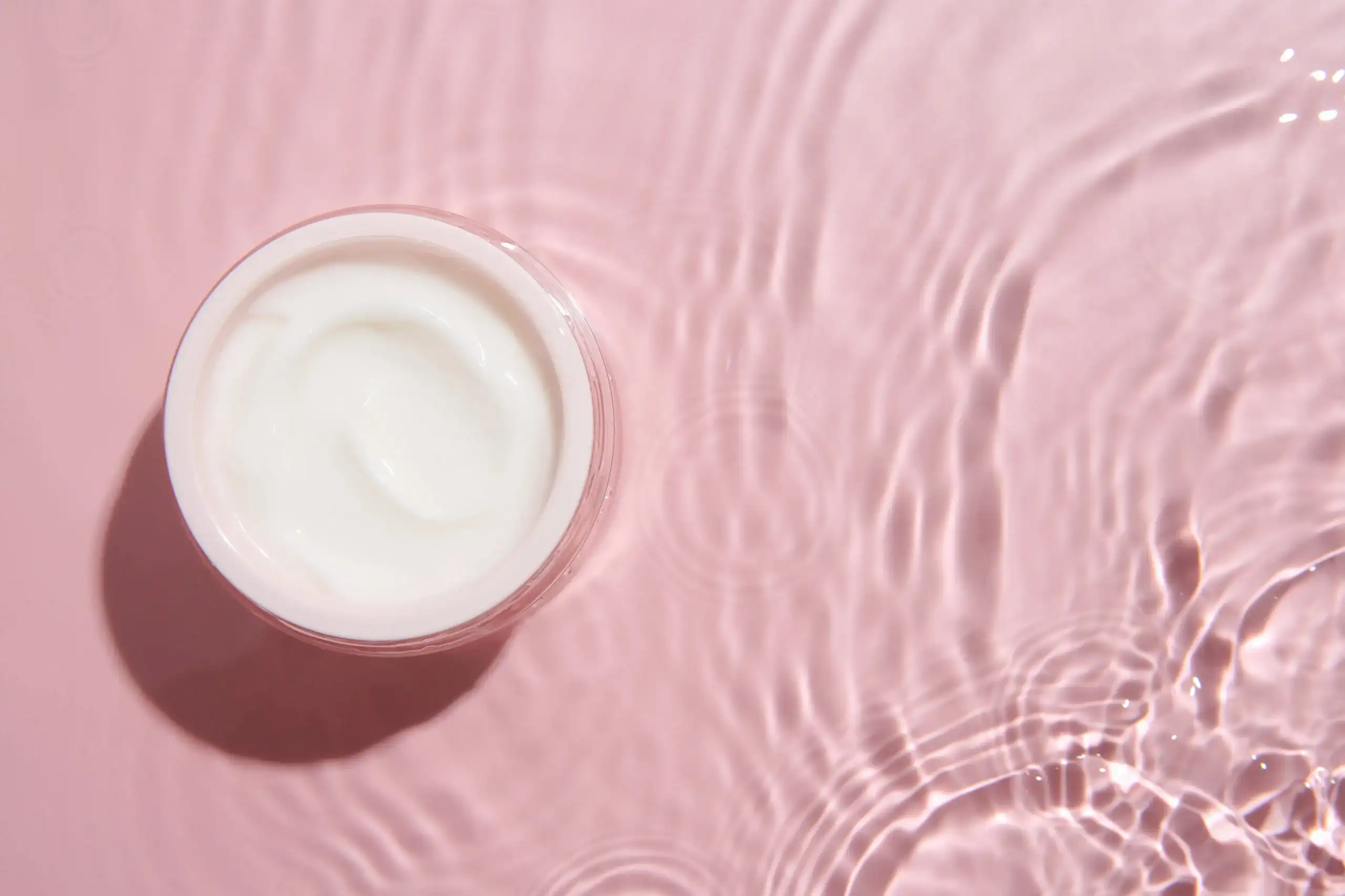 Moisturizer for dry skin on a pink surface with liquid around it.
